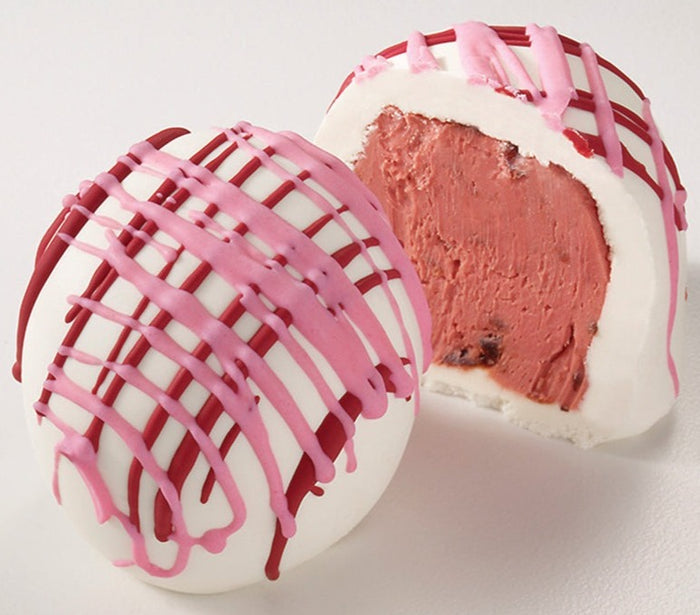 A photo of two cherry cheesecake truffles, one whole and one cut in half to show the filling, with white chocolate coating and pink drizzle.