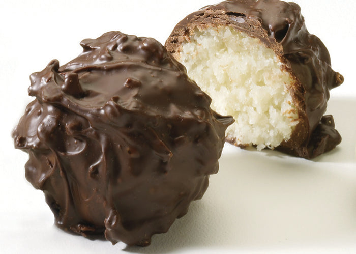Two chocolate covered coconut truffles on a white surface, one whole and one cut in half to show the creamy coconut center.