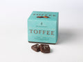 A box of Dark Chocolate Almond Sea Salt Toffee Squares with two squares placed in front, showing the chocolate and the almonds on top. Sweet Shop USA Mrs Weinstein's Toffee