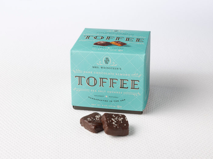 A box of Dark Chocolate Almond Sea Salt Toffee Squares with two squares placed in front, showing the chocolate and the almonds on top. Sweet Shop USA Mrs Weinstein's Toffee