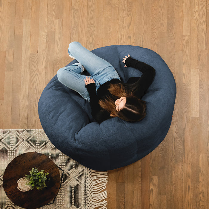 Person lying on a blue bean bag chair in a room with wooden flooring and a small table. #color_slate-blue-teddy-boucle