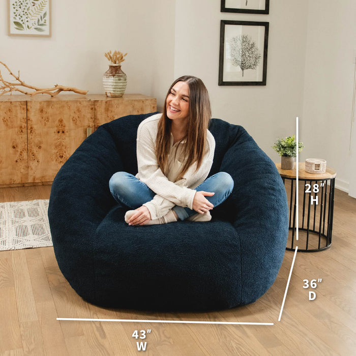 Woman sitting on a navy bean bag chair with dimensions displayed, in a home setting. #color_deep-blue-teddy-boucle