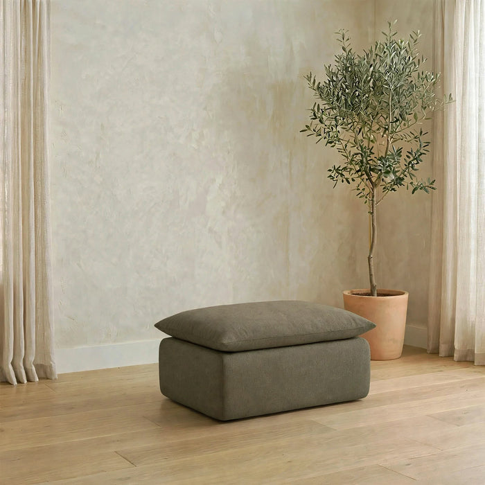 A rectangular ottoman with a cushioned lid in a muted taupe-gray fabric, placed on a light wood floor in a styled room with a potted olive tree and cream curtains.  #color_olive-green-terra-knit