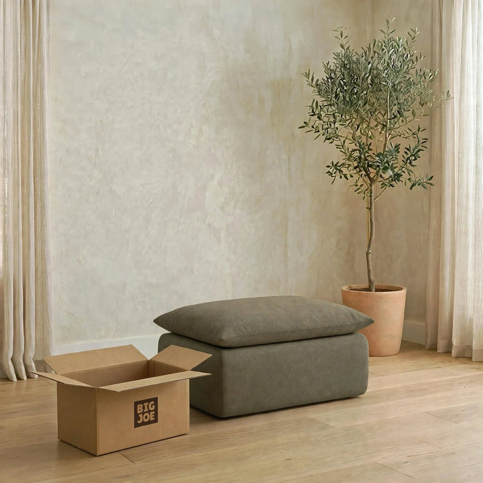 A gray upholstered ottoman with a matching rectangular cushion sits on a light wood floor in a styled room. A large open cardboard box with the visible brand 'BIG JOE' is on the left, and a potted olive tree stands against the wall on the right. #color_olive-green-terra-knit