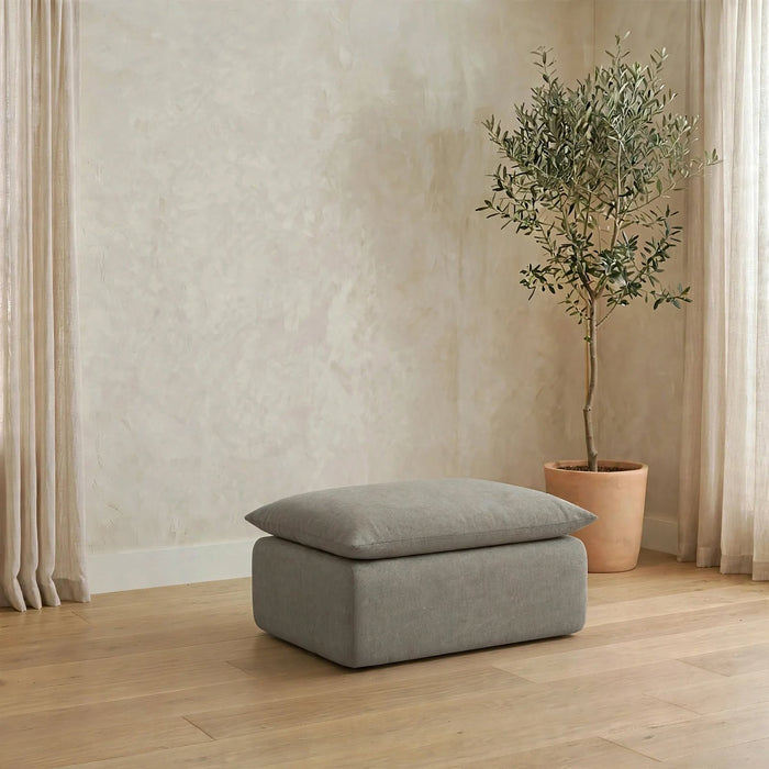 A rectangular ottoman with a matching cushion in a light gray fabric, placed on a light wood floor in a styled room with a potted olive tree and neutral walls. #color_misty-gray-terra-knit