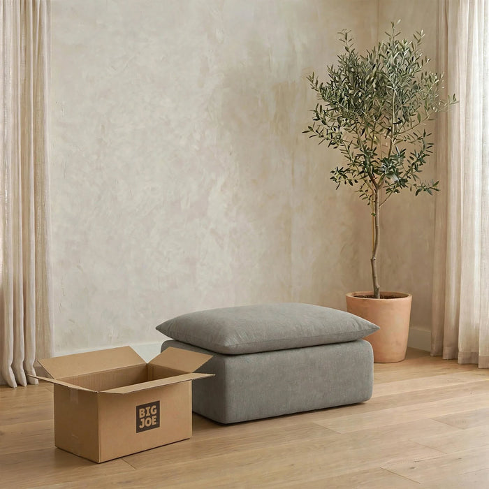 A gray upholstered ottoman or bench with two matching cushions sits on a light wood floor in a styled room. A cardboard box with the visible brand Big Joe is beside it, and a potted olive tree stands against a textured beige wall with sheer curtains. #color_misty-gray-terra-knit