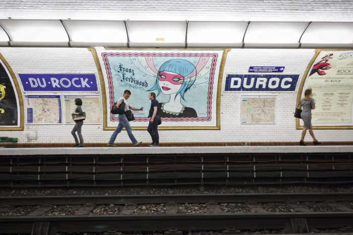 People walking in a subway station with a large mural of Tara McPherson's Franz Ferdinand gig poster on the wall.