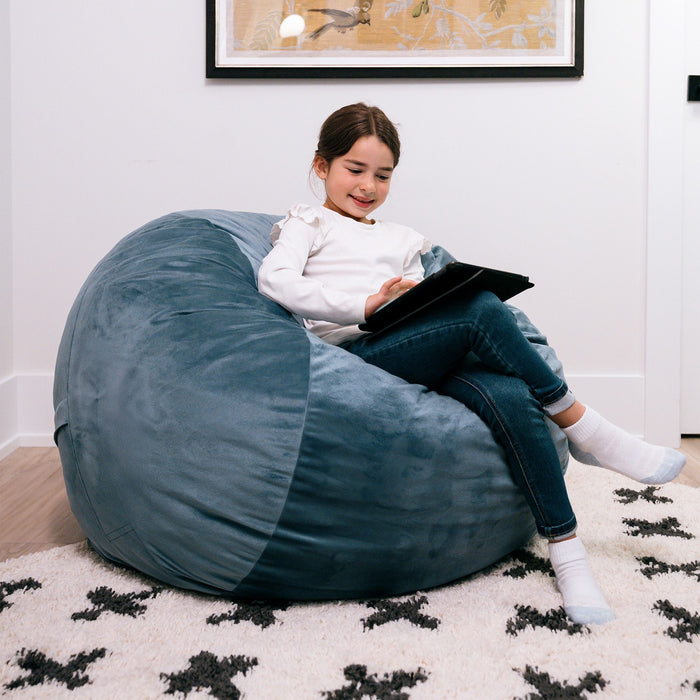 Child sitting on a large blue bean bag reading a book in a cozy room. #color_slate-blue-plush