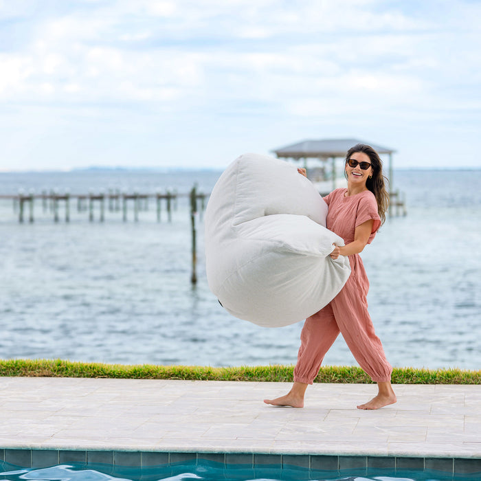Woman holding a luft bean bag chair by a pool with water and sky in the background #color_sunlit-ivory-outdoor-performance-linen