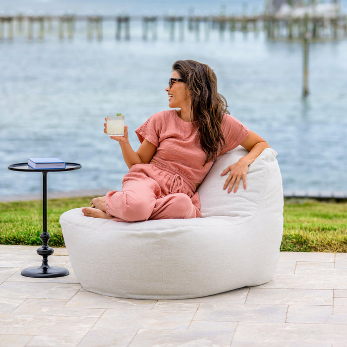 Woman sitting on a white bean bag chair by a waterfront, holding a drink. #color_sunlit-ivory-outdoor-performance-linen