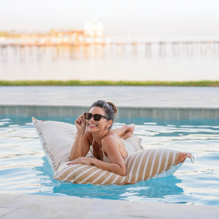 Woman lounging on a striped inflatable pool float in a pool. #color_sand-stripe-sunbrella-fabric