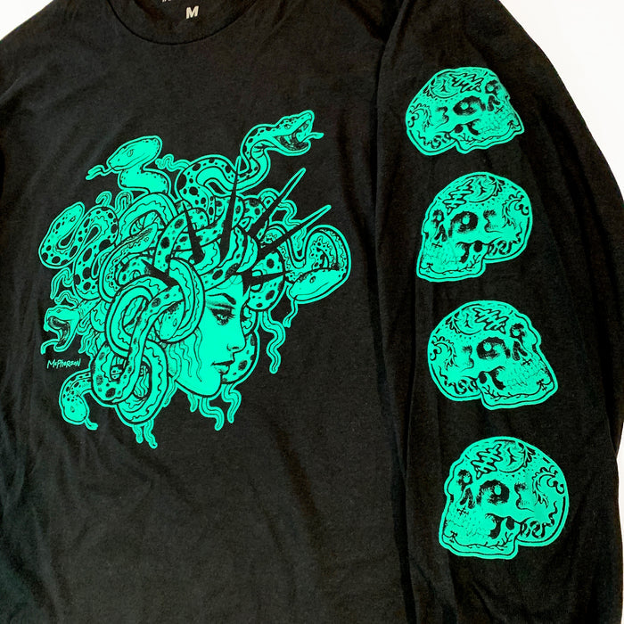 A black long-sleeve graphic T-shirt laid flat on a white background. The front features a large neon green illustration of Medusa with snakes and skulls, and the sleeves have three matching green skull graphics with stylized text.