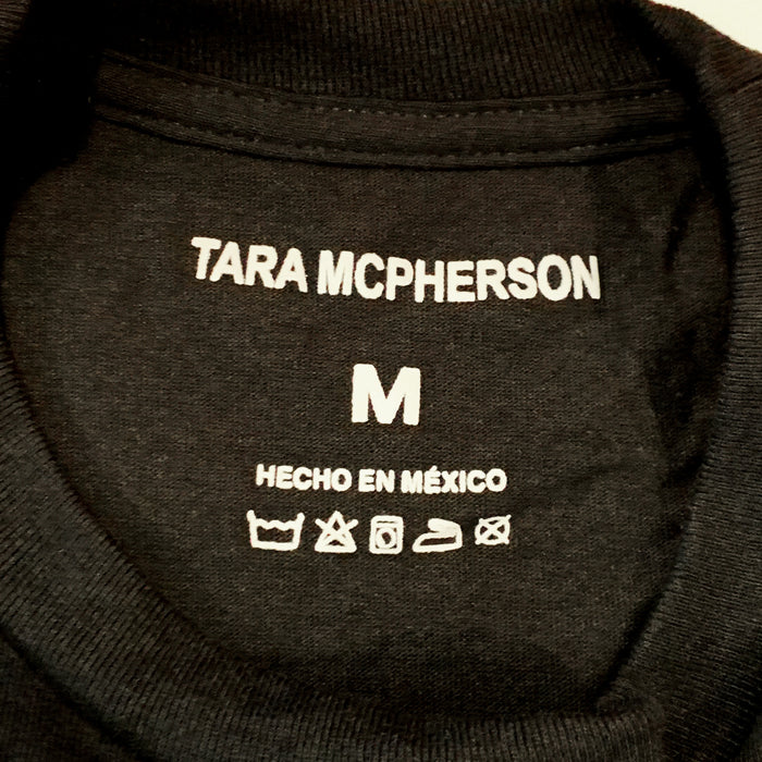 Close-up of the inside collar of a black crewneck sweatshirt, showing white printed text that reads 'TARA MCPHERSON', size 'M', 'HECHO EN MÉXICO', and care symbols.