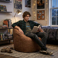 Person sitting on a bean bag chair reading a book in a cozy room with posters and shelves. #color_chestnut-vegan-suede