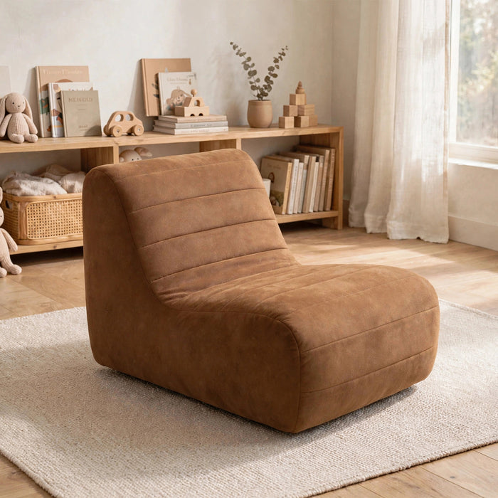A plush, wedge-shaped lounge chair in a warm camel brown faux suede material, styled on a light beige rug in a bright, modern nursery or playroom setting. #color_chestnut-vegan-suede
