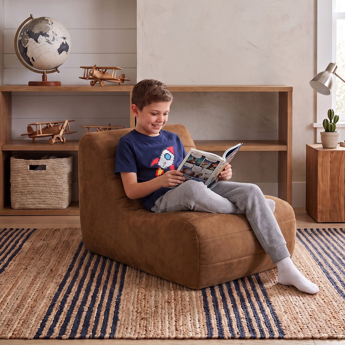 A young boy sits comfortably in a brown suede-like bean bag chair, reading a book in a cozy, well-lit room with a wooden bookshelf, globe, and striped rug. #color_chestnut-vegan-suede