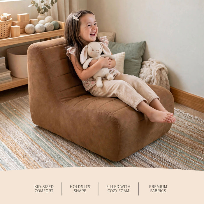 A young girl sits happily on a brown upholstered lounge chair in a styled nursery or playroom, holding a plush bunny toy. The scene includes a striped rug, wooden shelf, and decorative pillows in the background. #color_chestnut-vegan-suede