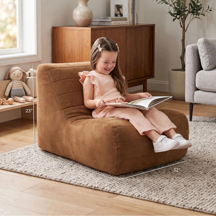 A young girl sits smiling in a brown upholstered armchair in a styled living room, reading a book. White dimension lines overlay the chair showing its 23 x 32 x 70 inch measurements. #color_chestnut-vegan-suede