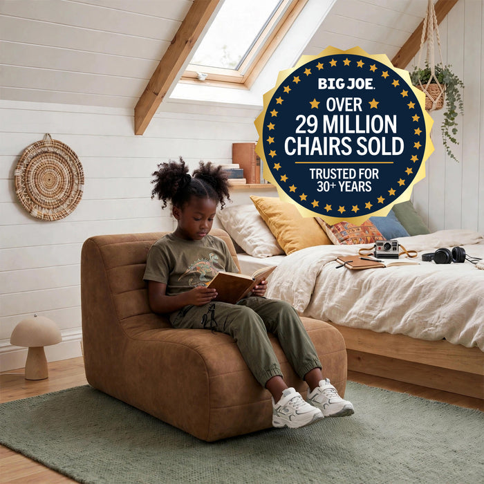 A child sits in a brown upholstered armchair reading in a bright bedroom with white wood walls, a bed, skylight, and green rug. A large circular badge reads 'BIG JOE OVER 29 MILLION CHAIRS SOLD TRUSTED FOR 30+ YEARS. #color_chestnut-vegan-suede