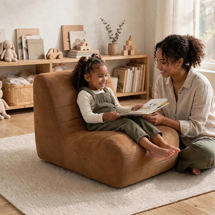 A woman and a young girl are reading a book in a cozy, warmly lit room. The girl is sitting in a plush, tan-brown upholstered lounge chair, while the woman kneels beside her on a light beige rug. #color_chestnut-vegan-suede