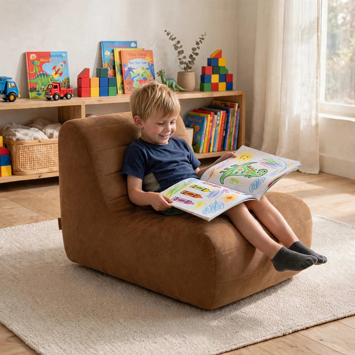 A young boy sits in a brown upholstered lounge chair reading a colorful book in a bright children's room with toy shelves and a window behind him. #color_chestnut-vegan-suede