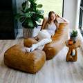 A woman relaxes on a large tan leather lounge chair with an attached ottoman in a bright, modern room. She is wearing a light gray tank top, white pants, and white socks. #color_copper-harbor-premium-vegan-leather