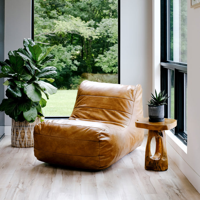 A tan leather chaise lounge chair positioned in a bright room with large windows, a green forest view, a potted fiddle-leaf fig plant, and a small wooden side table with a succulent. #color_copper-harbor-premium-vegan-leather