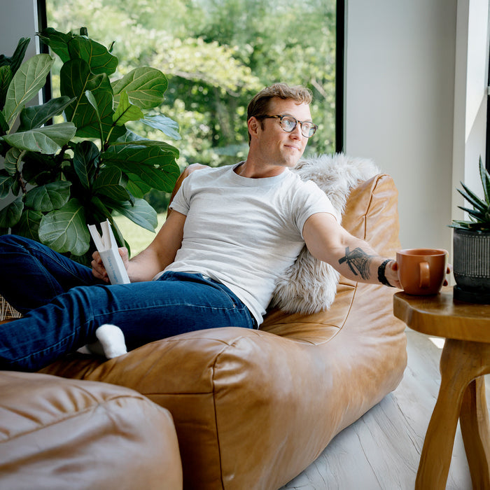 Man sitting on a brown leather sofa with a cup in a bright room with plants #color_copper-harbor-premium-vegan-leather