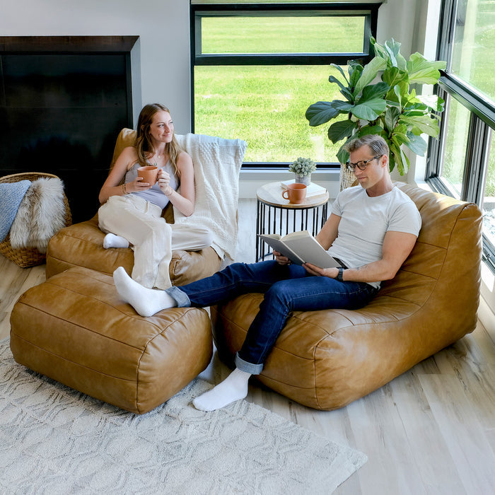 A man and a woman relax in oversized tan leather bean bag chairs in a bright, modern living room. The woman holds a mug and reads a book, while the man wears glasses and a white t-shirt. #color_copper-harbor-premium-vegan-leather