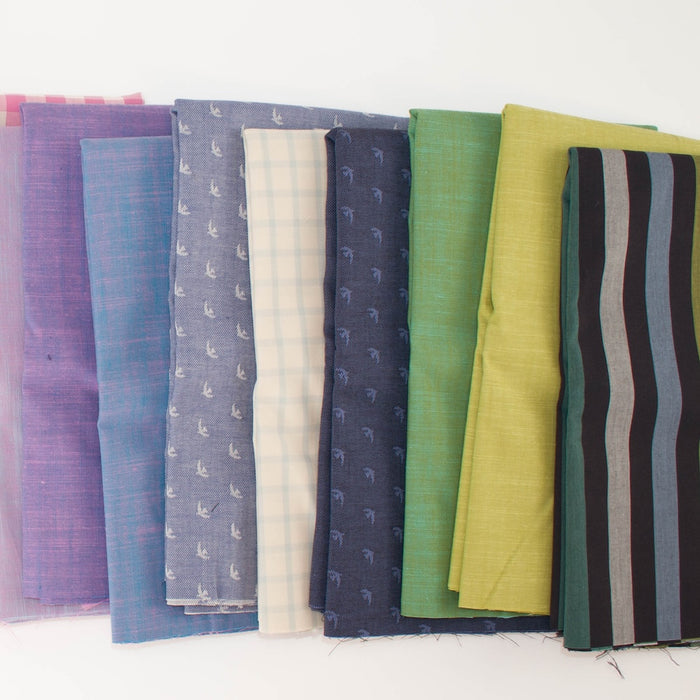 A row of folded fabric samples in multiple colors and patterns, including purple, blue, cream plaid, navy with small motifs, green, yellow-green, and black-and-gray stripes, arranged on a white background.