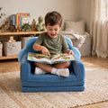 A young child sitting cross-legged in a small blue plush armchair, reading a colorful children's book in a softly lit playroom. #style_blue-dinosaur