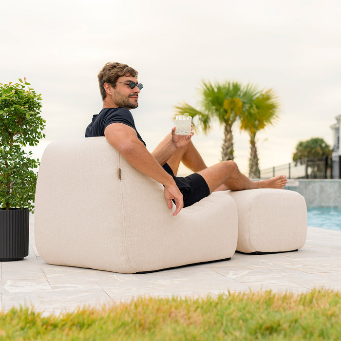 Man sitting on a beige outdoor chair by a pool with palm trees in the background #color_sunlit-cream-outdoor-performance-linen
