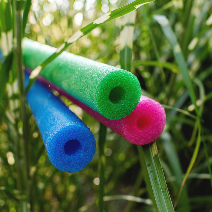 Three foam pool noodles in blue, green, and pink resting on tall green grass outdoors. #color_blue-green-pink