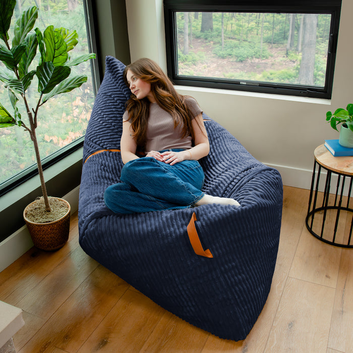 A woman reclines on a large navy blue ribbed bean bag chair in a bright room with wood floors, large windows, a potted plant, and a small side table. #color_midnight-blue-corded-plush