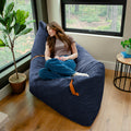 A woman reclines on a large navy blue ribbed bean bag chair in a bright room with wood floors, large windows, a potted plant, and a small side table. #color_midnight-blue-corded-plush