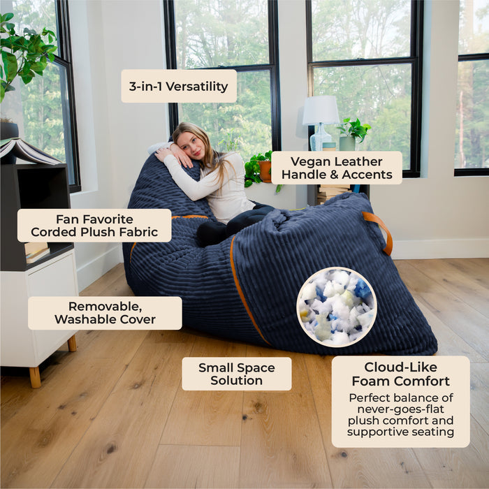 A woman reclines on a large ribbed bean bag chair in a bright living room. The bean bag is dark navy blue with orange trim and a circular inset showing the plush foam interior. #color_midnight-blue-corded-plush