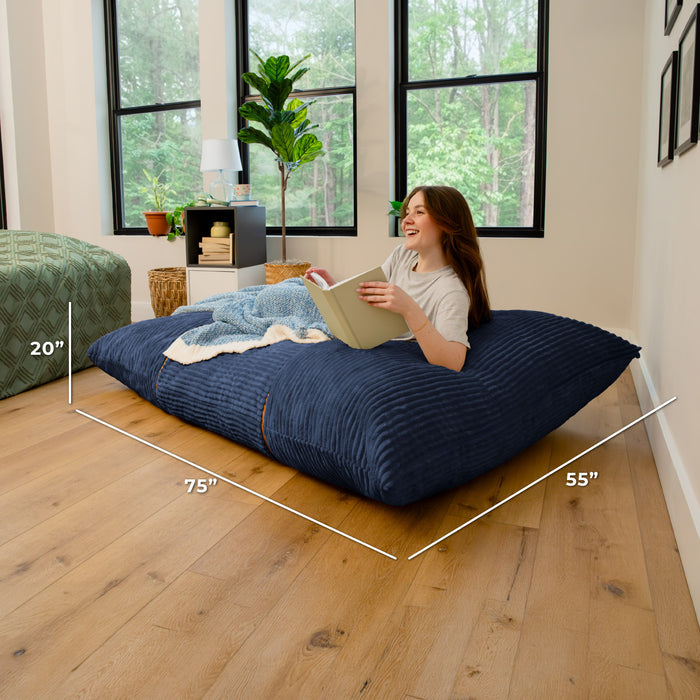 A woman reclines on a large navy blue ribbed floor cushion in a bright room with wood floors and large windows. White dimension lines overlay the cushion showing measurements of 75 inches by 55 inches, with a 20-inch height note. #color_midnight-blue-corded-plush