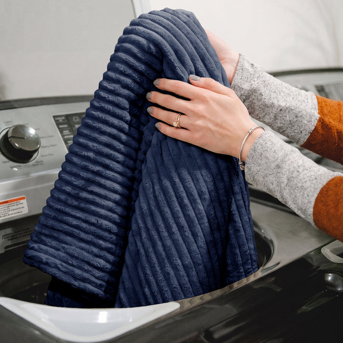 A person's hands and arms are shown holding a navy blue ribbed fleece blanket or towel in front of a washing machine. #color_midnight-blue-corded-plush