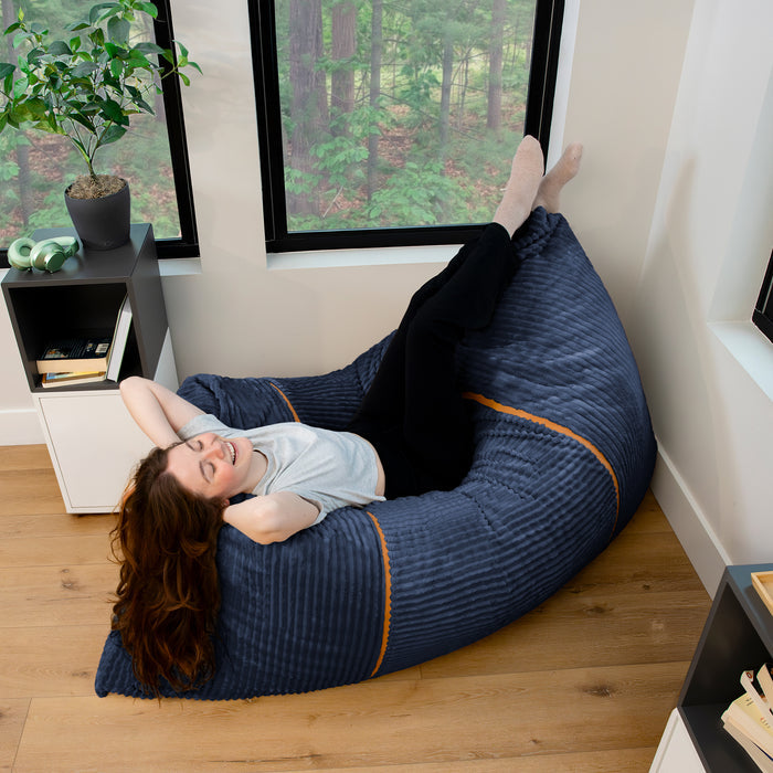 A woman relaxes on a large navy blue bean bag chair with orange piping, set in a modern room with wood floors and large windows. #color_midnight-blue-corded-plush
