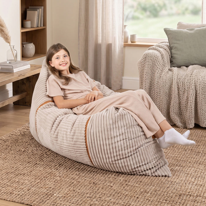 A young girl wearing beige pajamas sits comfortably on a large ribbed beige bean bag chair in a cozy, sunlit living room. #color_vanilla-latte-corded-plush