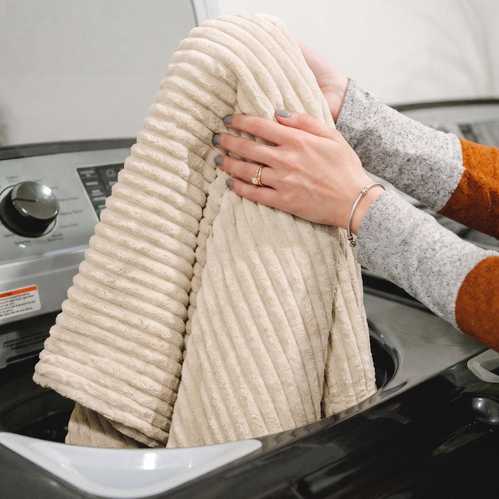 A person holding a folded ribbed cream corduroy blanket or towel over a washing machine. #color_vanilla-latte-corded-plush