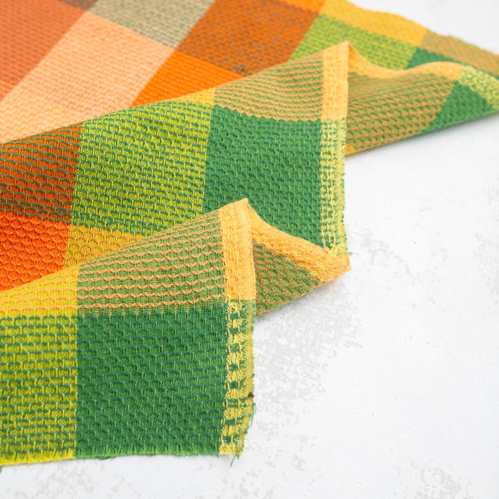A close-up of a woven fabric with a plaid pattern in green, yellow, orange, and beige, folded diagonally on a light gray textured surface.