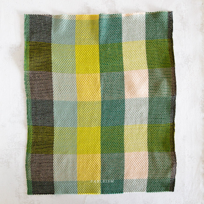 A square woven fabric swatch with a plaid pattern in shades of green, yellow, beige, and brown, laid flat on a textured off-white background.