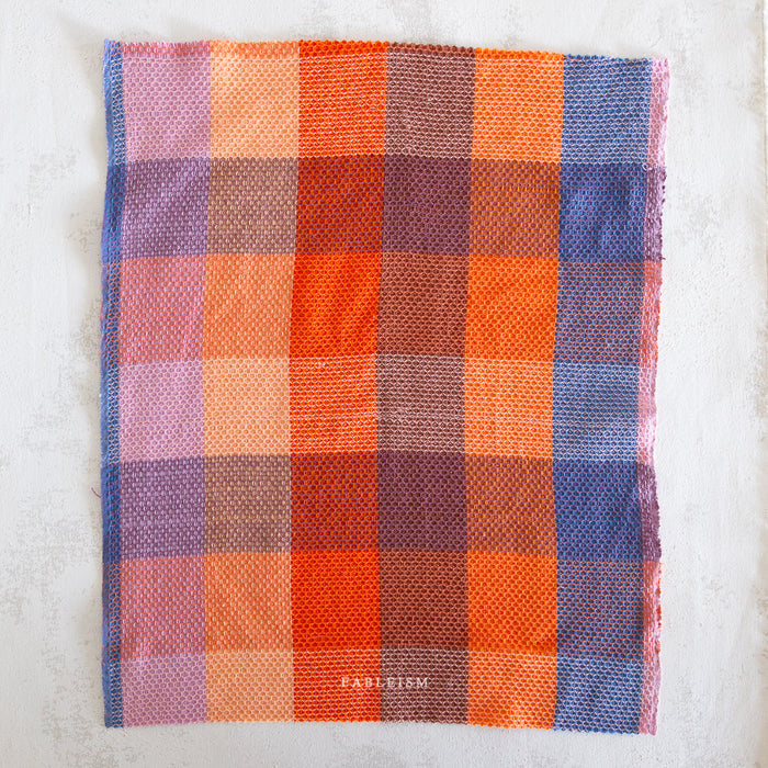 A square woven textile with a multicolor plaid pattern in shades of orange, pink, purple, and blue, laid flat on a light textured background. The brand name 'Fableism' is printed in white near the bottom center.